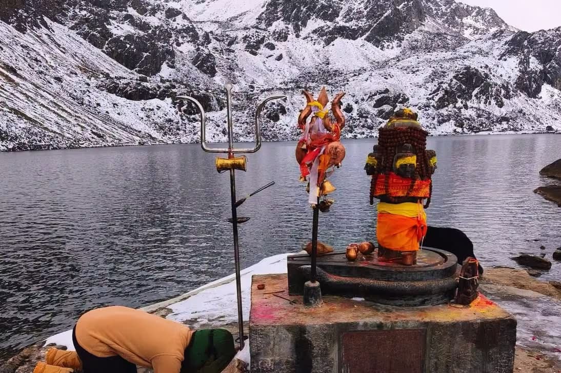 A black stone image of Shiva wrapped in rudraksha malas and an orange cloth on a plinth at the lake's edge, a trident and a bell-hung staff beside it, snow on the ground and on the peaks across the dark water