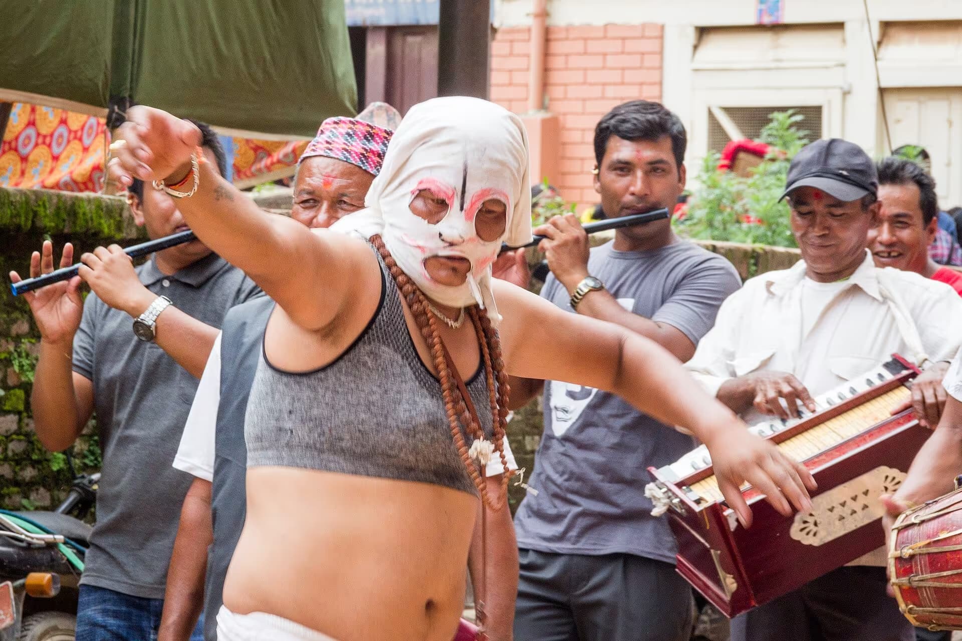 A costumed performer in white face paint dancing ahead of a band with flutes and a harmonium