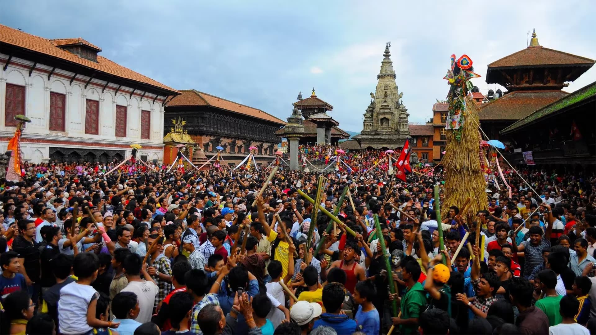 A tall straw and bamboo effigy raised over a packed Bhaktapur Durbar Square, sticks held up in the crowd
