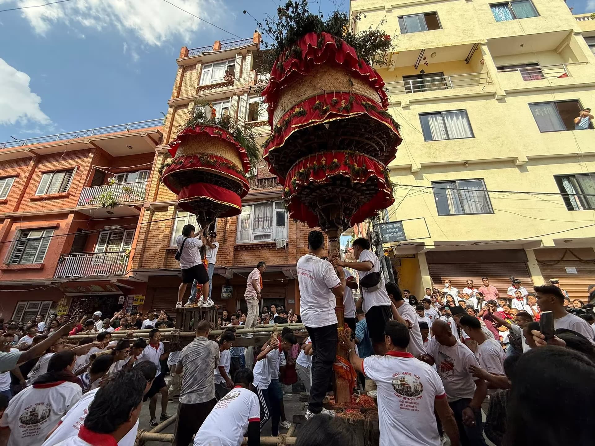 Two tiered khats draped in red cloth and greenery, widest at the top, held aloft on poles above a Hadigaun street packed with bearers in white Chokde Narayan Jatra T-shirts