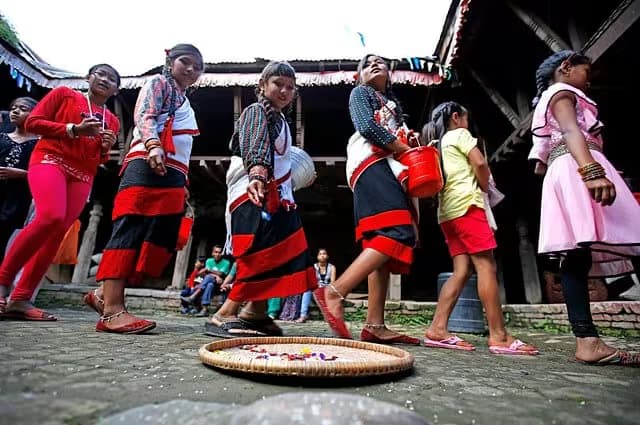 Girls in Newar dress carrying offering pots past a woven tray of flowers in a Patan courtyard during Matya