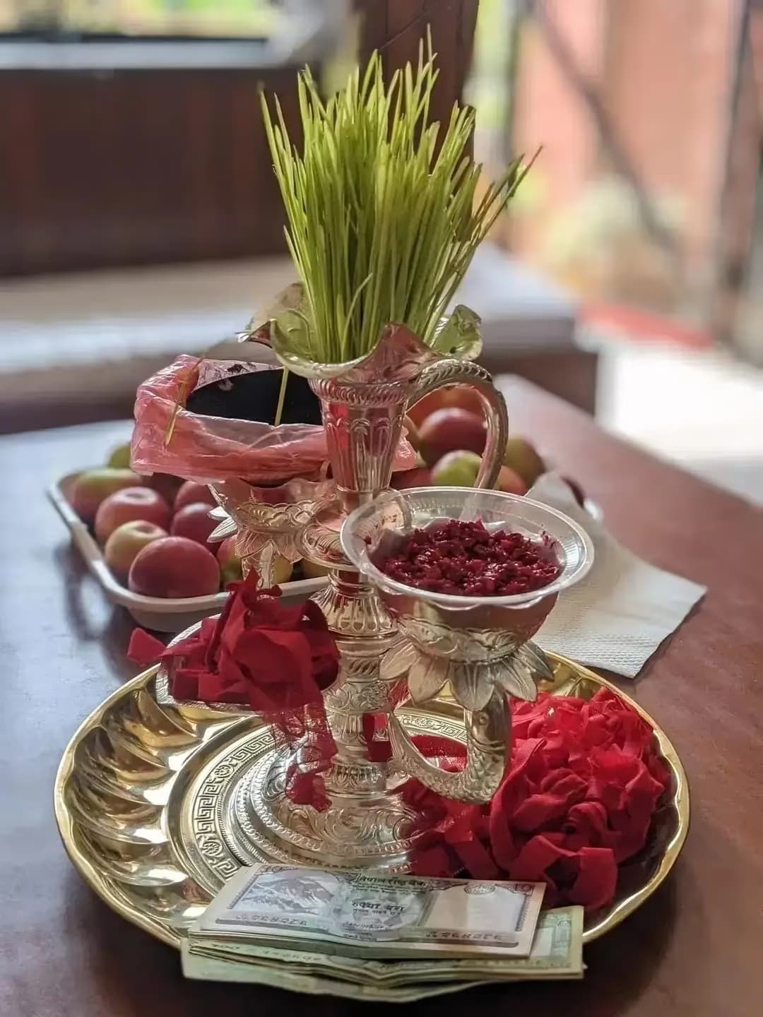 A Dashain tika plate holding jamara shoots, red tika powder, apples and banknotes