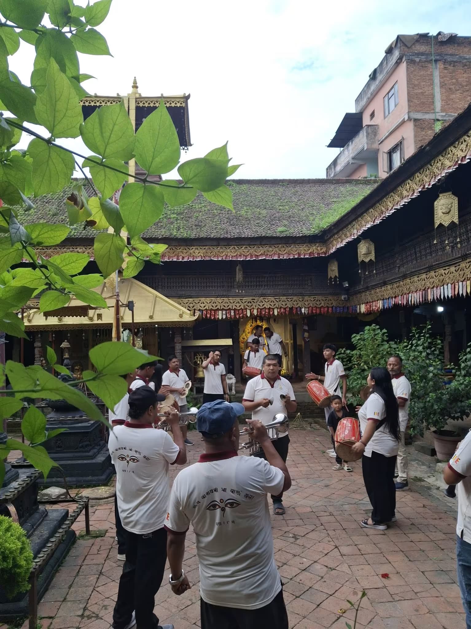 Musicians with drums and cymbals playing in the brick courtyard of a bahal, the tiled monastery roofs rising around them