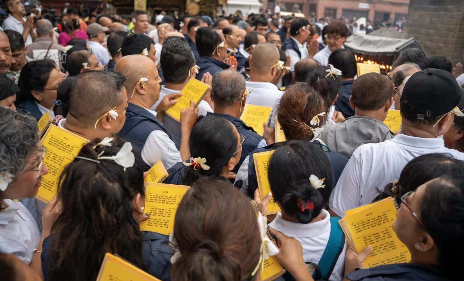 A dense crowd of devotees, flowers tucked behind their ears, reading aloud from yellow scripture booklets
