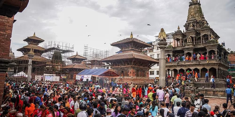 A dense crowd crossing Patan Durbar Square towards the Krishna Mandir on the right, tiered temple roofs and scaffolding along the far side, incense smoke rising from the middle of the square